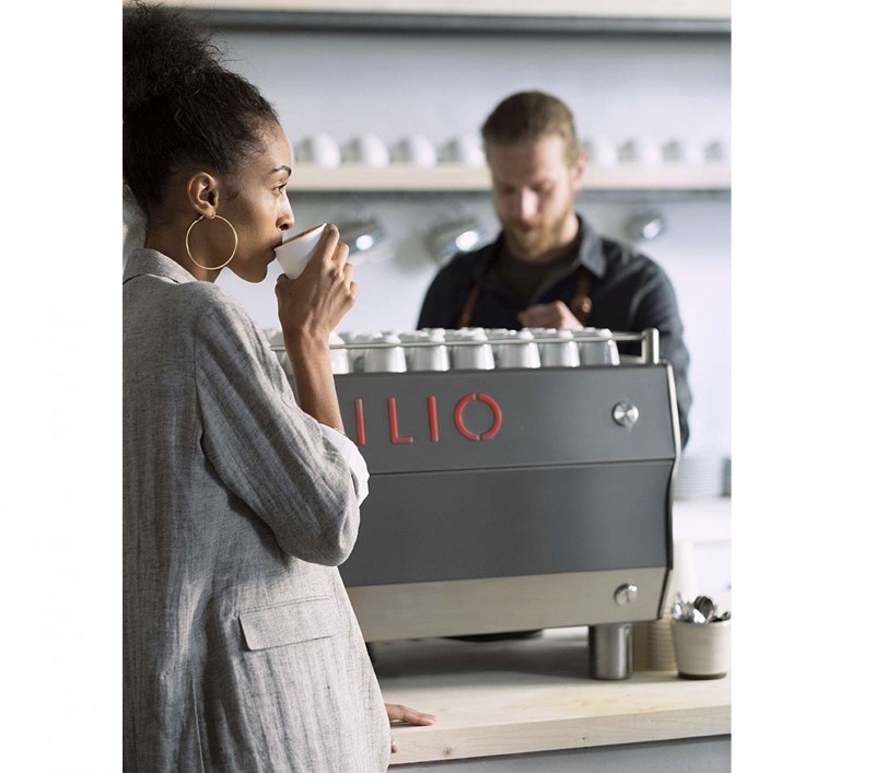 This image is a front-side view of the Rancilio Specialty RS1 espresso machine showcase picture with barista and customer with drink.